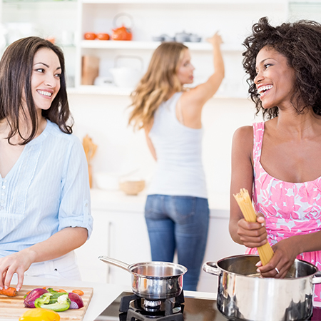 Group of three women cooking a meal together in a gas stove kitchen setting.